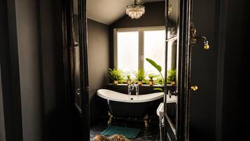 Ein Badezimmer mit freistehender Badewanne, einem Fenster mit Pflanzen, einer Chandelieber und einer Tür mit Messingklinke.