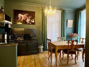 Une cuisine avec des meubles de rangement aux couleurs sombres, une table à manger en bois et des chaises. On y trouve un lustre, un tableau accroché au mur et une fenêtre avec des rideaux.