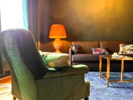 Una poltrona verde con un cuscino, un divano con una coperta fantasia, un tavolino da caffè in legno e una lampada in una stanza con tappeto blu.