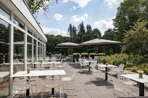 An outdoor patio with white wicker chairs and tables, large umbrellas, and a view of trees and greenery. - Explore Scenic Trails & Relax at This Garden Retreat with Free Parking Included (Amersfoort)