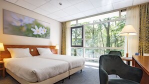 A hotel room with a double bed, a desk, a chair, a large window with a view of trees, and a painting on the wall.