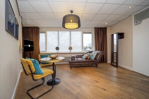 Ein modernes Wohnzimmer mit Holzboden, einem runden Esstisch, zwei Sofas, einem Fernseher und einem großen Fenster mit Vorhängen.