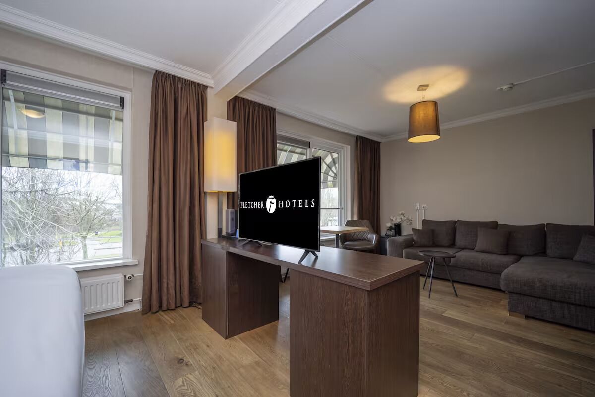 A modern hotel room with a large flat-screen TV, a wooden desk, a sofa, and a lamp.