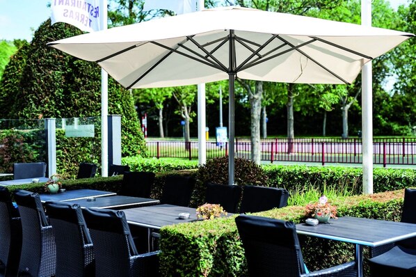 Un espace extérieur aménagé avec des tables et des chaises, protégé par un grand parasol, et entouré de verdure et d’une clôture.