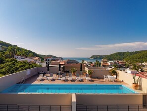 Ein Pool auf dem Dach mit Liegestühlen und Sonnenschirmen mit Blick auf eine Küstenstadt und das Meer.