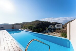 A swimming pool with a wooden deck, surrounded by buildings and hills. - 2BR Condo Steps to Beach | Costta A Santa Cruz (Huatulco)