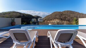 Ein Poolbereich mit zwei weißen Liegestühlen, einer Holzterrasse und Blick auf einen Berg und Gebäude im Hintergrund.