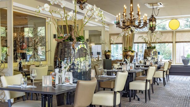 Un restaurant avec des tables dressées pour le service, de grands vases décoratifs remplis de fleurs et un lustre.