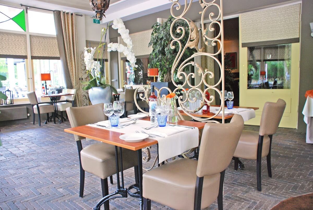 Un comedor con suelo de ladrillo, sillas color beige, una mesa de madera preparada para cuatro comensales y una partición decorativa de metal.