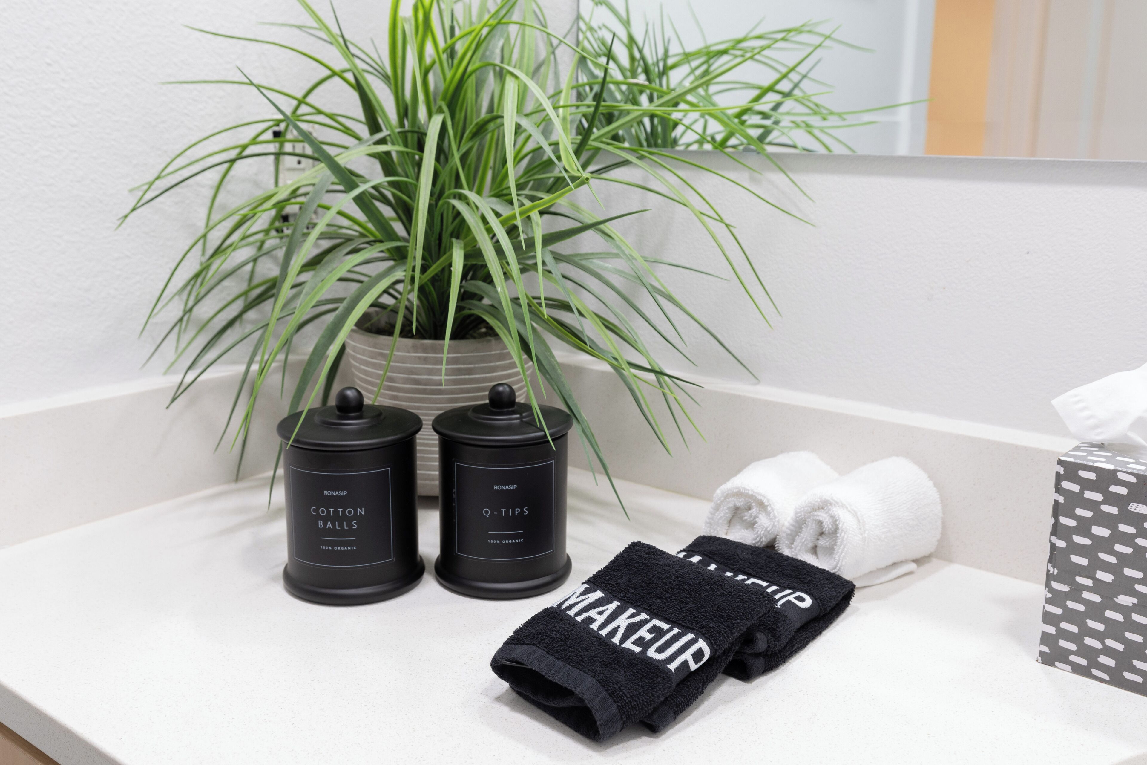Een badkamer met een plant, twee zwarte bakjes met de tekst 'Cotton Balls', een zwarte handdoek met 'MAKEUP' erop en een witte handdoek.