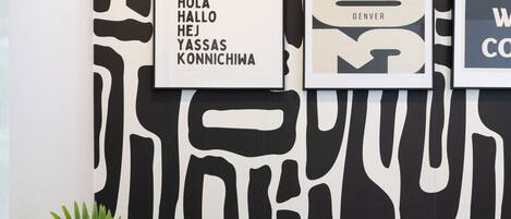 A wall with abstract black and white patterns, a framed sign with multilingual greetings, and a decorative plant in a textured vase.