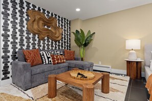 Ein modernes Wohnzimmer mit einem grauen Sofa, gemusterten Kissen, einem Holztisch, einer großen Pflanze und einer Lampe auf einem Beistelltisch.
