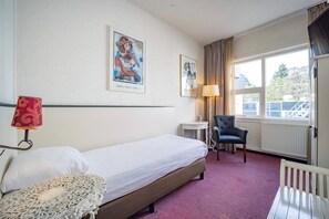 Ein Hotelzimmer mit einem Bett, einem Sessel, einem kleinen Tisch, einer Lampe und zwei gerahmten Bildern an der Wand.