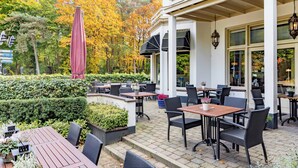 Un espace extérieur aménagé en terrasse, avec des tables et des chaises, entouré d’une haie et d’arbres aux feuilles d’automne.