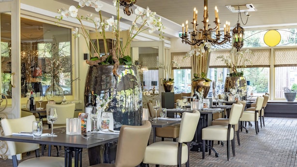 Un restaurant avec des tables dressées pour le service, de grands vases décoratifs remplis de fleurs et un lustre.