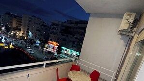 A balcony with a round table and red chairs, overlooking a cityscape at night.