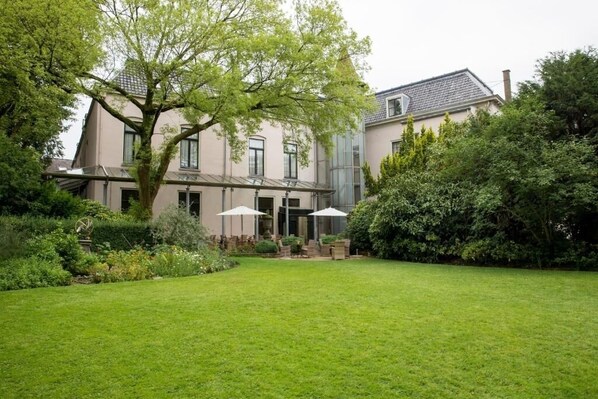 A two-story house with a large front lawn, a tree, and a patio area with umbrellas.