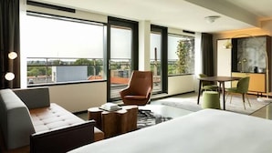 Habitación de hotel moderna con una cama grande, un sofá, una silla, una mesa de comedor y vistas de la ciudad a través de amplias ventanas.