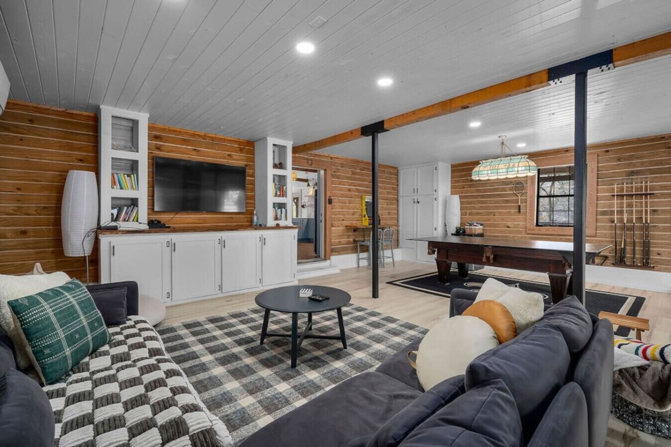 A modern living room with a sofa, a coffee table, and a TV. There is a kitchen area with cabinets and a dining table.