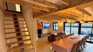 Eine Holztreppe führt zu einem Erker, dort befinden sich ein Esstisch mit Stühlen, eine Couch und ein Fernseher in einem Raum mit großen Fenstern.