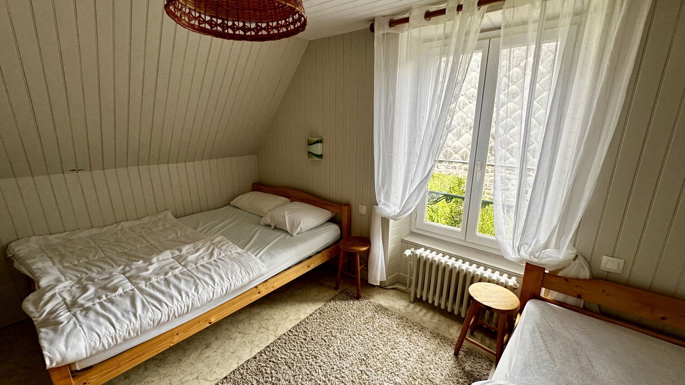A bedroom with a single bed, a small stool, a radiator, and a window with sheer curtains.