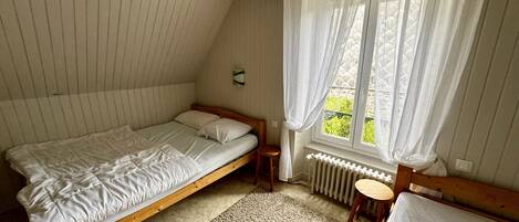 A bedroom with a single bed, a small stool, a radiator, and a window with sheer curtains.