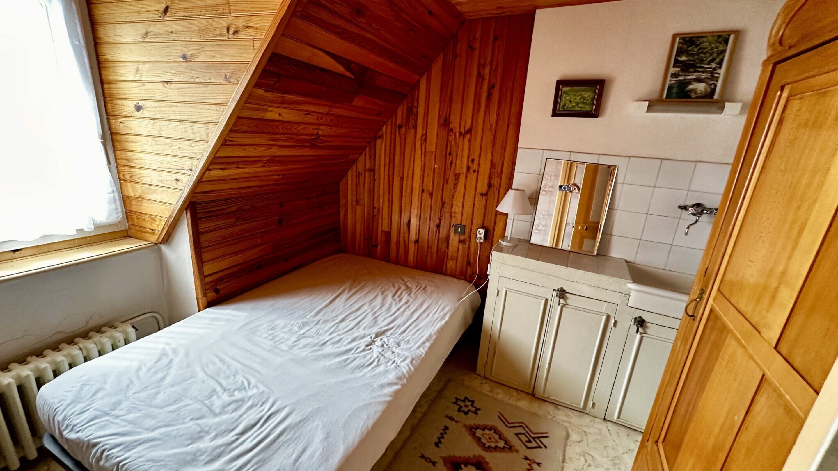 Una stanza piccola e accogliente con soffitto spiovente, un letto singolo, una piccola cassettiera e una finestra.