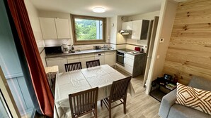 A compact kitchen with white cabinets, a dining table with four chairs, a microwave, a coffee maker, and a window with a view of greenery. - Le Capucin 79 - Le Mont Dore (Mont-Dore)