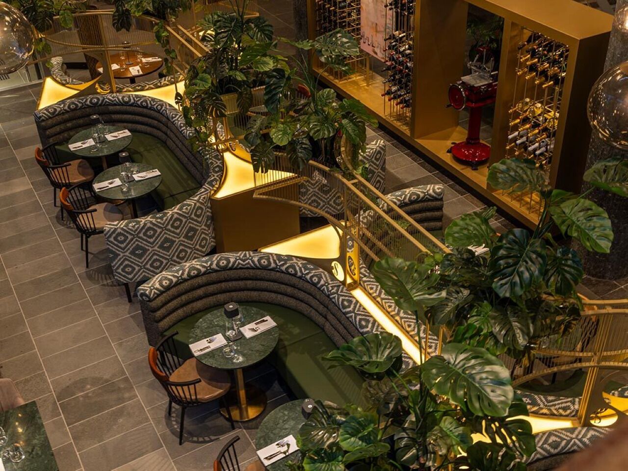 Un restaurant avec des tables rondes et des sièges rembourrés, entouré de grandes plantes.
