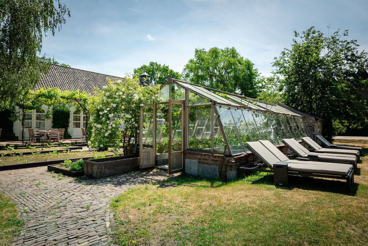 Een kas met glazen dak, omgeven door een tuin met een metselpad en lounge stoelen.