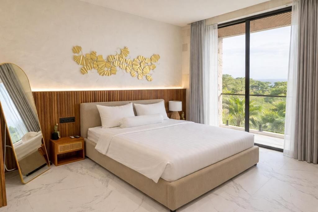 A hotel room with a large bed, a bedside table, a nightstand, a wall-mounted mirror, and a view of greenery through the window.