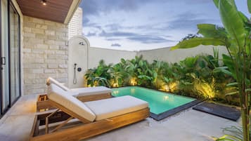 A poolside area with a lounge chair and a small pool, surrounded by lush greenery and a stone wall.