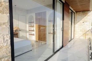 A modern bedroom with a large bed, a glass door leading to a balcony, and a stone wall. - White Residence (KUTA SELATAN)