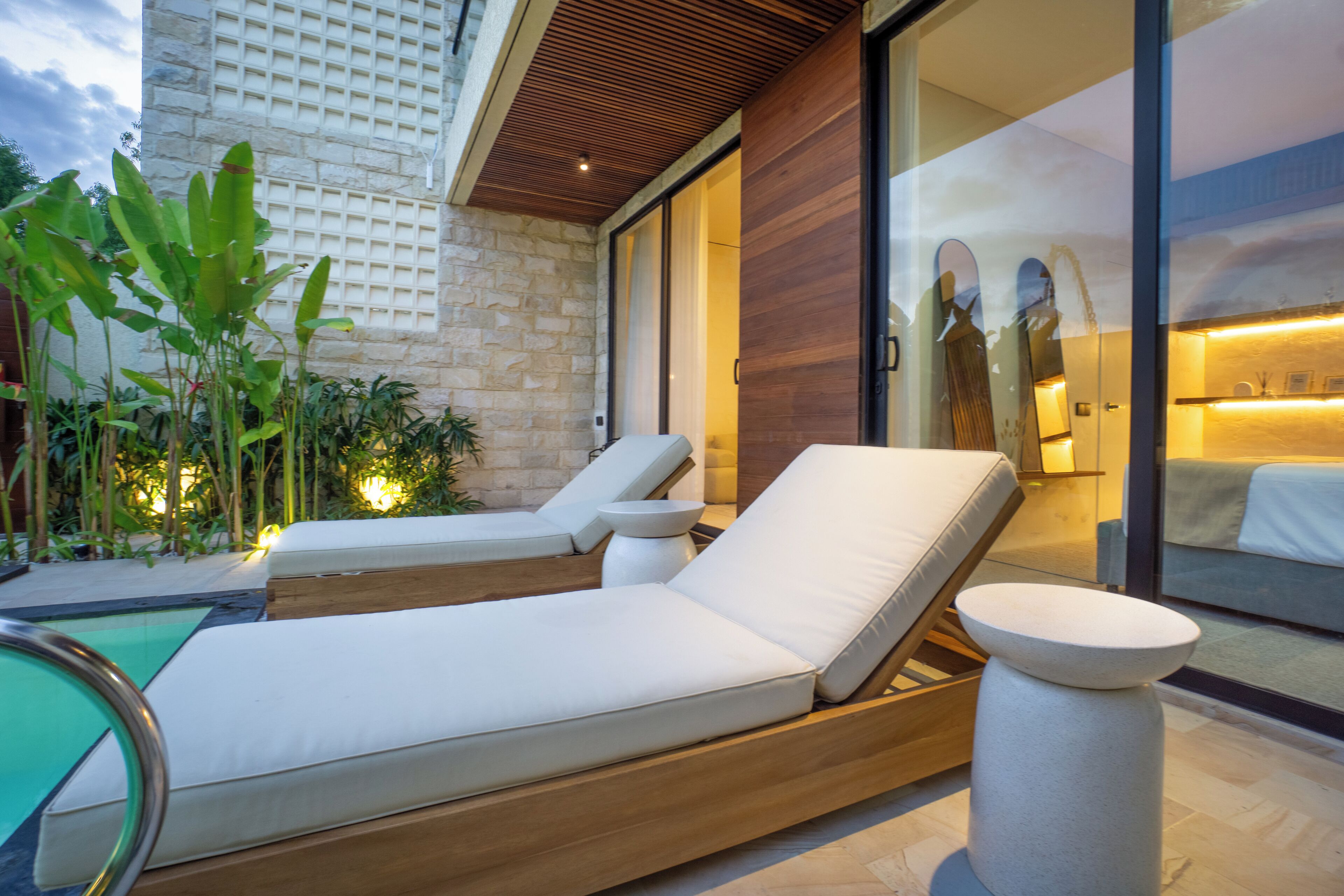 A modern outdoor lounge area with two sunbeds, a pool, and a stone wall.