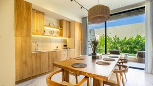 A modern kitchen with wooden cabinetry, a large dining table, and a view of lush greenery outside. - White Residence (KUTA SELATAN)