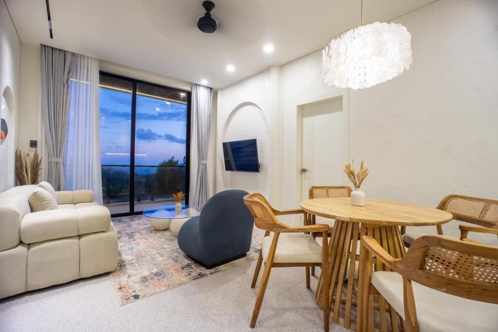 A modern living room with a sofa, a round wooden table, and chairs. There is a large window with curtains, a flat-screen TV mounted on the wall, and a decorative ceiling light.