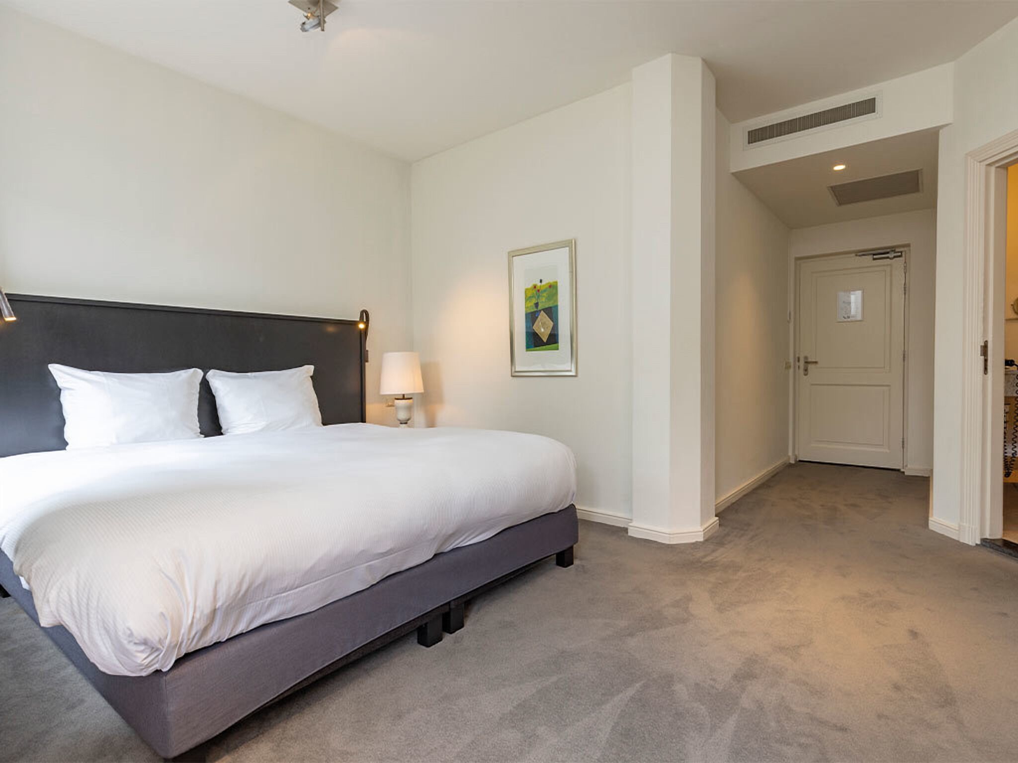 Quarto de hotel com uma cama grande, uma mesa de cabeceira com abajur e um quadro pendurado na parede.