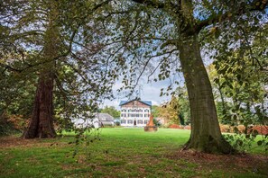 Een groot, traditioneel gebouw met een symmetrische gevel, omgeven door een verzorgd gazon en volwassen bomen.