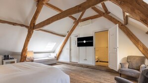 A hotel room with a sloped ceiling, wooden beams, a bed, a desk, a chair, and a TV screen displaying the hotel name. - Ideal Location! Free Parking, Onsite Dining! Close to Jochumhof Botanical Garden (Baarlo)