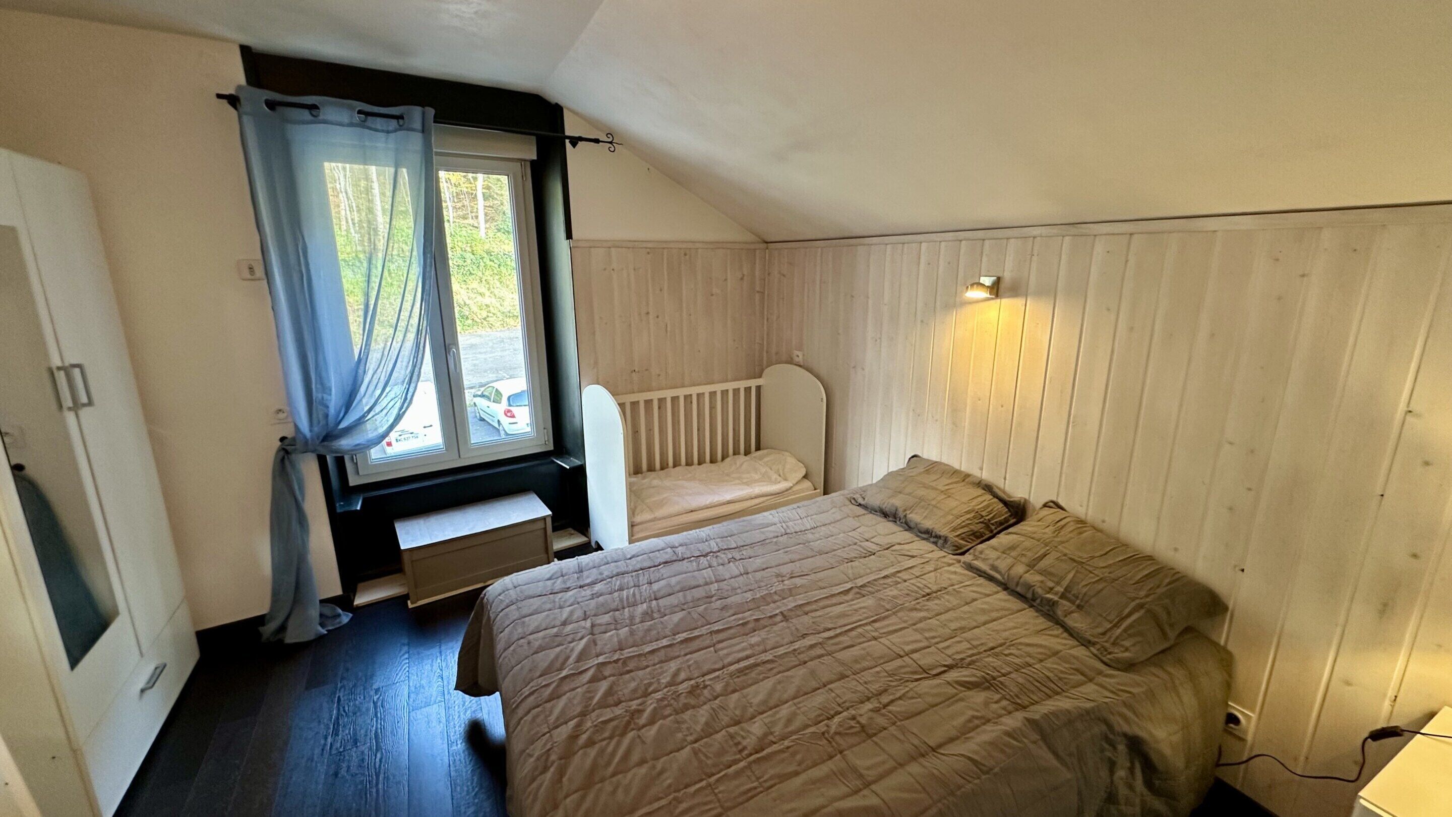 Une petite chambre avec un lit simple, un plancher en bois, une fenêtre avec des rideaux et un luminaire fixé au mur.