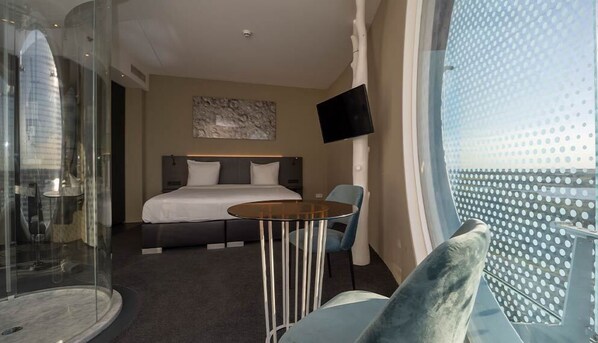 Una camera d'albergo con un letto, un tavolino, una sedia, una TV e un'ampia finestra vista un edificio.