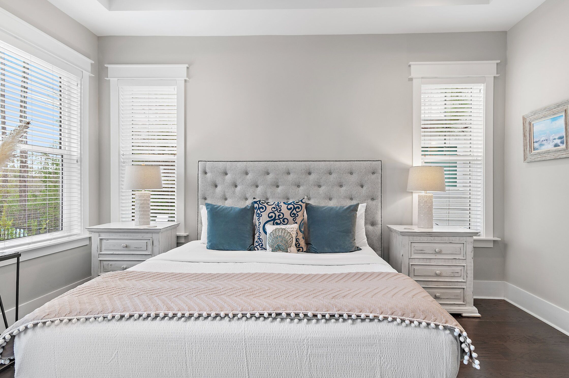 A bedroom with a tufted headboard, a large bed with a patterned bedspread, two bedside tables with lamps, and a window with blinds.