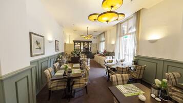 Een restaurantgedeelte van een hotel met meerdere tafels en stoelen, een bar op de achtergrond en hangende lampen.