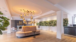 Un salon moderne avec un canapé rond, une petite table ronde et un lustre original. On y trouve une cheminée, une plante fixée au mur et, au fond, un coin cuisine.