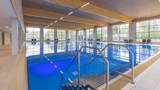 Une piscine intérieure à l’eau d’un bleu cristallin, avec des poutres en bois au plafond et de grandes fenêtres.