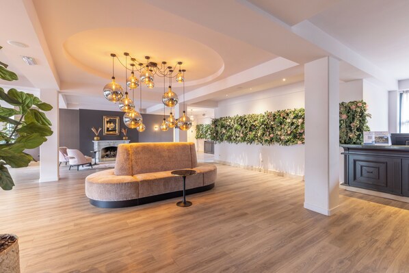 A modern living room with a round sofa, a small round table, and a unique chandelier. There is a fireplace, a wall-mounted plant, and a kitchen area in the background.