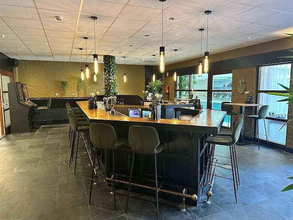A modern bar area with a long counter, multiple bar stools, pendant lighting, and a view of the outdoors through large windows.
