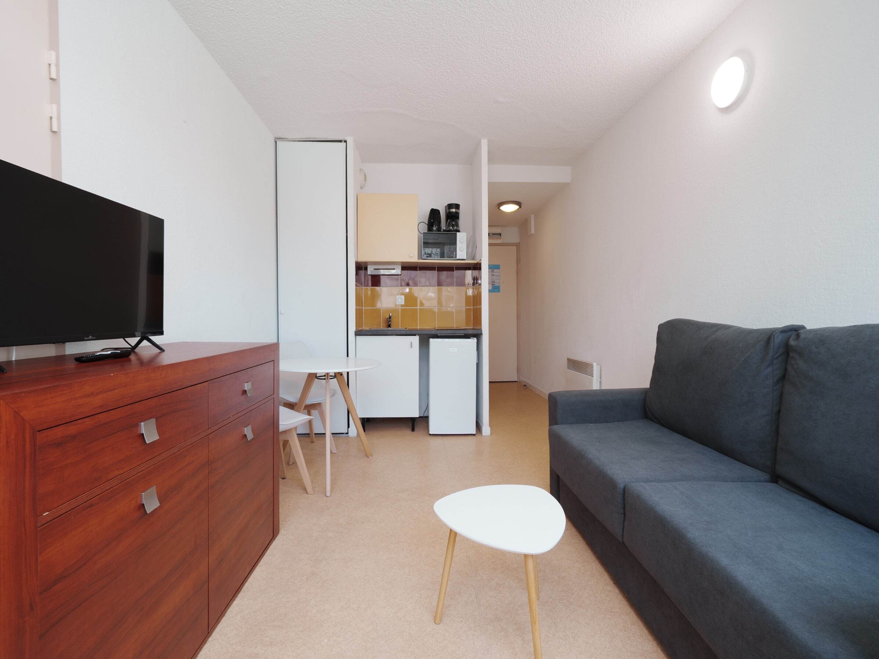 A compact living space with a TV on a wooden dresser, a gray sofa, a small white table, and a kitchenette in the background.