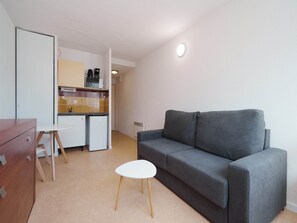 A compact living space with a grey sofa, a small white table, a kitchenette with a microwave, and a wooden cabinet. - Scenic Escape by the Village rim (Agde)