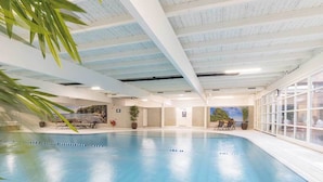 Una piscina interior con agua azul cristalina, rodeada de suelo y paredes con baldosas. Hay sillas de descanso y plantas en macetas alrededor de la piscina.